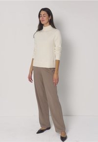 Frau steht mit geschlossenen Augen, trägt einen weißen Rollkragenpullover, lockere braune Hose und schwarze spitze Schuhe vor einem einfarbigen Hintergrund.