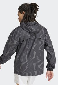 Graue wasserfeste Jacke mit Kapuze, ausgestattet mit einem wellenförmigen Muster und reflektierenden Akzenten. Lange Ärmel mit elastischen Bündchen und Reißverschlussbelüftungen.