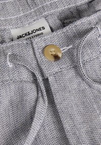 Gros plan sur un tissu de pantalon tissé gris clair, bouton doré, passants de ceinture et étiquette Jack & Jones visible à l'intérieur de la ceinture.