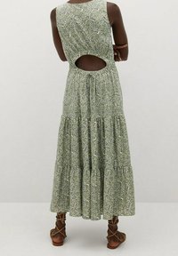 Robe maxi fleurie verte avec une coupe à volants, un détail dos découpé et un lien ajustable. Portée avec des sandales marron à lacets.