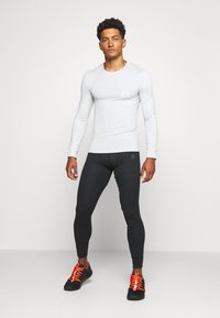 Vaaleanharmaa pitkähihaista paitaa, jossa on logo, mustat tiukat leggingsit ja mustat urheilukengät, joissa on kirkkaat oranssit nauhat, seisomassa valkoista taustaa vasten.