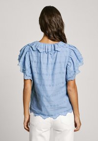 Blusa azul claro con mangas acampanadas, escote con forma de concha, detalles de ojal y tela texturizada. El dobladillo presenta un borde en forma de concha.