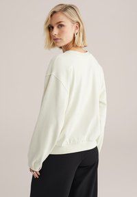 Hanorac crem deschis cu umeri căzuți, manșete striate și un guler rotund simplu, asortat cu pantaloni negri. Material moale, texturat.