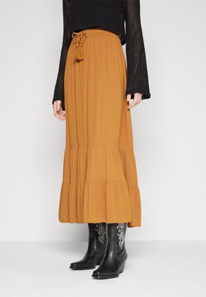 Kobieta nosząca musztardową, warstwową spódnicę maxi z paskiem z frędzlami, czarny, szydełkowany top z długim rękawem oraz czarne botki w stylu western z haftem.