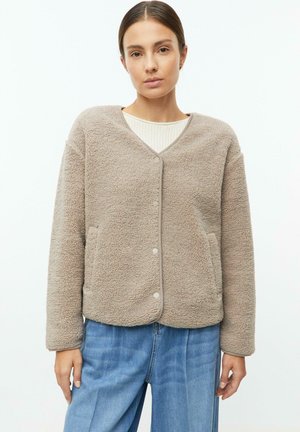 Frau trägt eine beige, geknöpfte Sherpa-Jacke über einem hellen Pullover und blauen Jeans und steht vor einem schlichten weißen Hintergrund.