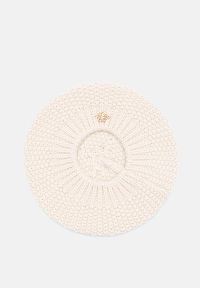 Béret rond tricoté couleur crème avec centre côtelé et petit emblème circulaire doré sur le dessus, présenté sur fond blanc.