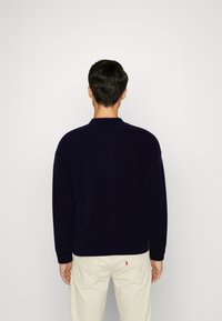 Navy sweater with a rounded collar and long sleeves, made of textured fabric, styled with a relaxed fit and paired with light-colored pants.
