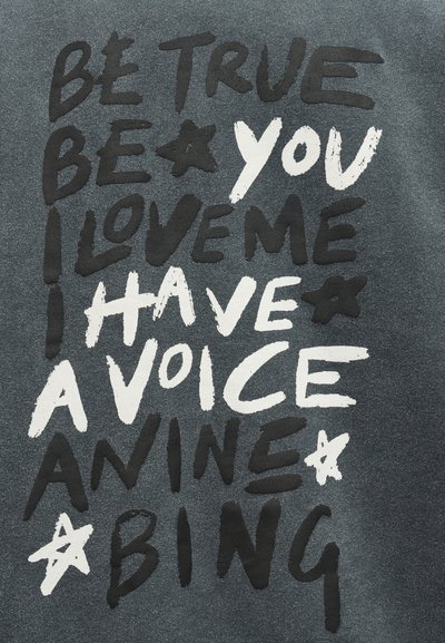 Juoda ir balta spalvų raidės ant tamsios audinio fono su užrašu „Būk tikras, būk savimi, myliu save, turiu balsą, animė, bing“.