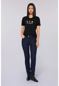 Černé tričko s krátkým rukávem s logem a grafickou potiskem, v kombinaci s tmavě modrými přiléhavými džíny. Model stojí vzpřímeně v černých kotníkových botách.