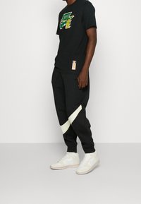 Czarna bawełniana koszulka z zielonym i żółtym logo Nike; czarne dresy z kremowym ukośnym paskiem; białe sneakersy z szarymi akcentami.