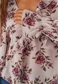 Blommig blus med trumpetärmar, tillverkad av ett lätt genomskinligt tyg i ljusrosa, prydd med mörkrosa och gröna ros-mönster.