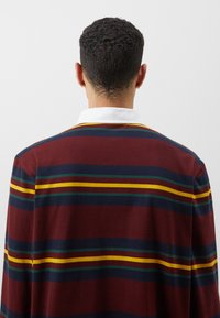 Bordeaux gestreiftes Polohemd mit weißem Kragen. Verfügt über horizontale Streifen in Gelb, Grün und Blau. Glatte Textur, lässiges Design.