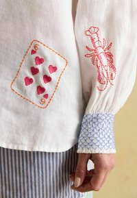 Weißes Leinenhemd mit rosa Herz- und orangefarbener Kartenstickerei an den Ärmeln, mit einem blauen Rüschensaum. Gestreifter Stoff darunter.