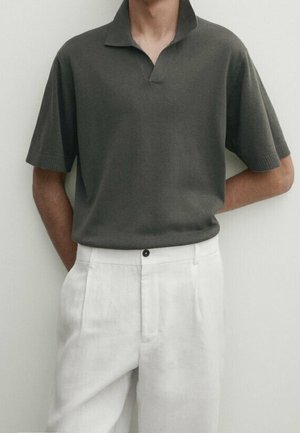 Homme portant un polo en maille gris foncé à manches courtes, rentré dans un pantalon blanc plissé taille haute avec un bouton noir.