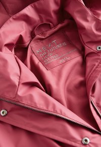 Rózsaszín, sima szövetkabát belső része, fém címkével, amelyen az áll: „NEXT THE FUTURE IS NOW”, cipzárral és patentokkal felszerelve.