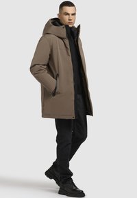 Bruine geïsoleerde jas met een capuchon, zwarte rits, zijzakken en een licht taps toelopende vorm, gecombineerd met zwarte jeans en schoenen.