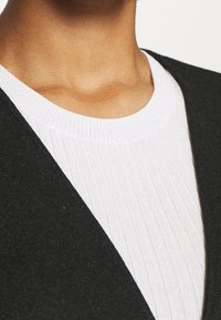 Černý vlněný cardigan s překrývajícím se límcem nad bílým žebrovaným svetrem, zdůrazňující kontrastní textury a barvy na švu oděvu.
