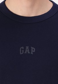 Chemise à manches longues côtelée bleu marine avec un col rond et un subtil logo "GAP" gris sur la poitrine. Tissu texturé avec une coupe ajustée.