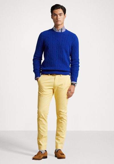 Blå kabelstrikket sweater, gule chinos, brune loafers, synlige striber på skjorteærmerne. Glat stof med teksturerede mønstre og skræddersyet pasform.