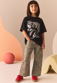 T-shirt nera oversize con grafica dei Rolling Stones, abbinata a pantaloni larghi beige con stampa leopardata e sneakers rosse, su uno sfondo pastello morbido.