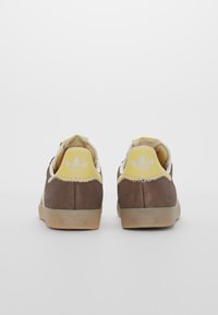 Zapatillas de ante marrón con detalles en amarillo, lengüeta blanca y cordones. Suela de goma texturizada y logotipo en el talón. Vista trasera de los zapatos.