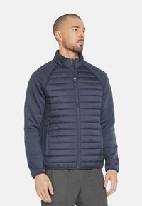 Marineblauwe pufferjack met horizontaal gestikt ontwerp, contrasterende fleece mouwen, hoge kraag en volledige ritssluiting aan de voorkant. Twee zijzakken zijn inbegrepen.