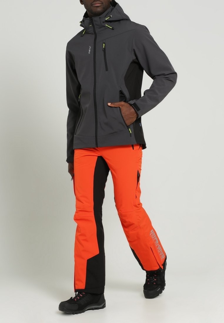 Homme portant une veste à capuche gris foncé et un pantalon de plein air orange vif et noir avec des chaussures de randonnée noires, debout devant un fond uni.