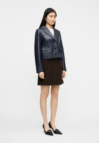 Femme debout portant une veste en cuir bleu marine boutonnée, une jupe en daim marron avec poches avant, des talons noirs à bouts pointus, sur fond blanc uni.