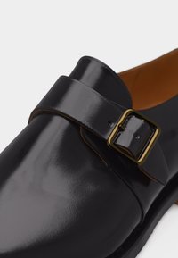 Mocassin en cuir noir poli avec une boucle rectangulaire dorée et une bande traversant le dessus, laissant entrevoir une partie de la doublure intérieure couleur beige.