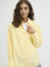 Gul knappet skjorte med lange ermer, laget av glatt stoff. Har klassisk krage og subtile sømdetaljer. Brukt over en hvit topp.