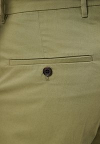 Olivgrön tyg med en slät yta. Har en bakficka med en mörkbrun knapp och subtila sömmar.