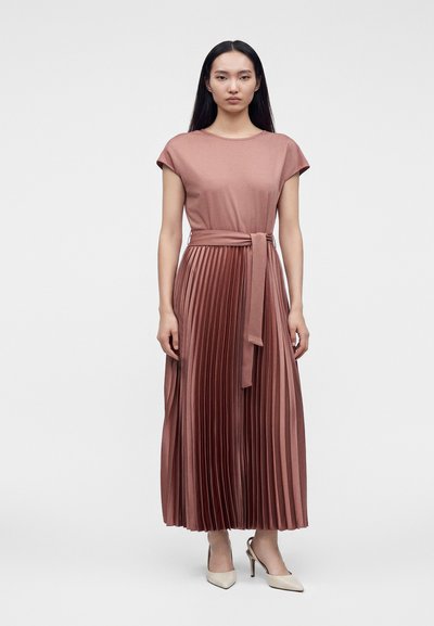 Femme debout portant une robe midi plissée rose poudré à manches courtes avec une ceinture assortie, associée à des talons pointus crème.