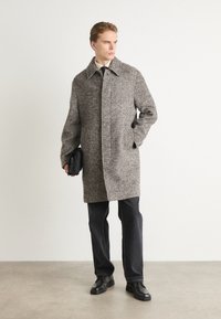 Palton gri cu model houndstooth, la guler, cu nasturi pe față și buzunare pe lateral. Asortat cu pantaloni negri și încălțăminte, ținând o geantă neagră.