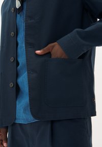 Jachetă ușoară de culoare albastru marin, cu închidere cu nasturi, buzunar pe piept și textură netedă. Purtați-o peste o cămașă din denim, evidențiind un design stratificat.