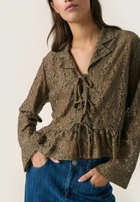 Olive green lace blouse with a peplum hem, featuring a tie closure and long sleeves, textured floral pattern, worn over blue denim jeans.