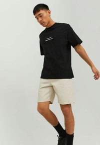 T-shirt nera in cotone con maniche corte e testo bianco, abbinata a pantaloni corti beige. Modello che indossa calze nere e scarpe nere.