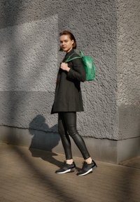 Svart dubbelknäppt kappa, åtsittande svarta leggings, svarta träningsskor och en grön läderryggsäck. Mjuka texturer och slimmad design.