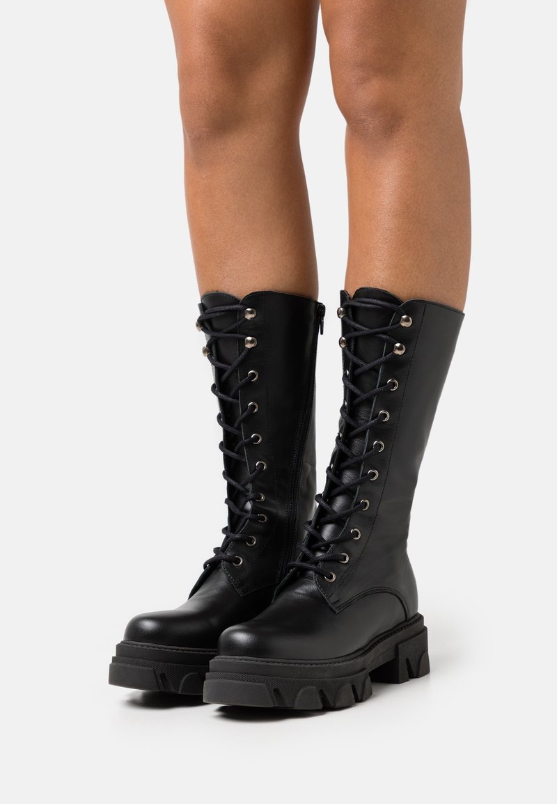 Stivali alti in pelle nera con lacci frontali, occhielli in argento e suole grosse e testurizzate. Finitura liscia con dettaglio a collarino ondulato.