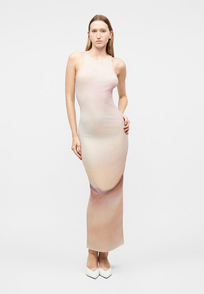 Γυναίκα με μακριά ίσια μαλλιά, φορούν αμάνικο, εφαρμοστό μακρύ φόρεμα σε διαβάθμιση από μπεζ σε ανοιχτό ροζ και λευκά μυτερά παπούτσια.