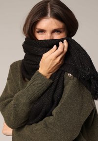 Écharpe noire texturée avec un motif en chevrons et des bords frangés, portée sur un pull vert. Tissu doux, apparence épaisse et confortable.