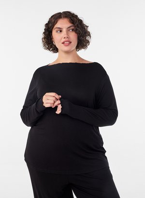 Donna con capelli ricci che indossa una maglia nera a maniche lunghe, guarda di lato, mani giunte vicino al petto, su uno sfondo uniforme.