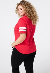 T-shirt rouge à manches courtes avec des rayures horizontales blanches sur les manches, en tissu lisse, associé à un pantalon noir.