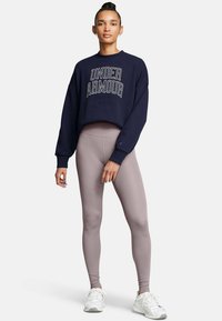 Tummansininen huppari, jossa on "Under Armour" -teksti; löysä malli. Vaaleat legginsit; tiukka, sileä pinta. Valkoiset urheilukengät nauhoilla.