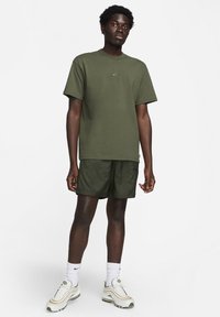 Vīrietis stāvē, ģērbies olīvu zaļā Nike t-kreklā un šortos, baltās Nike zeķēs un baltās čībās uz vienkrāsu gaiša fona.