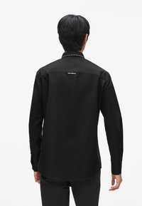 Fekete, hosszú ujjú póló sima textúrával, which studded gallérral és egy kis márkajelzéssel a hátsó felső részen. Klasszikus dizájn és illeszkedés.