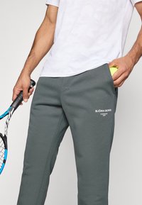 Calças de moletom cinzentas com uma textura suave, apresentando um logótipo e bolsos laterais, combinadas com uma t-shirt branca com padrão e uma raquete de ténis.