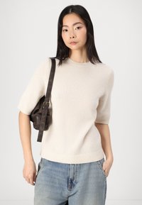 Helles beiges Strickpullover mit kurzen Ärmeln und geripptem Saum, kombiniert mit hellblauen lässigen Jeans und einer kleinen braunen Ledertasche für die Schulter.