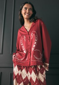 Rotes Hemd mit Knopfverschluss, das auf der Vorderseite weiße Schwanendesigns zeigt, kombiniert mit gemusterten Hosen in Beige und Rot mit Schwanmotiven.