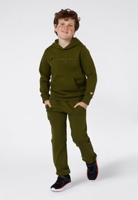 Felpa con cappuccio in cotone verde oliva con logo "Champion" sul petto, abbinata a pantaloni della tuta verdi oliva. Sneakers nere con accenti rossi e verdi.
