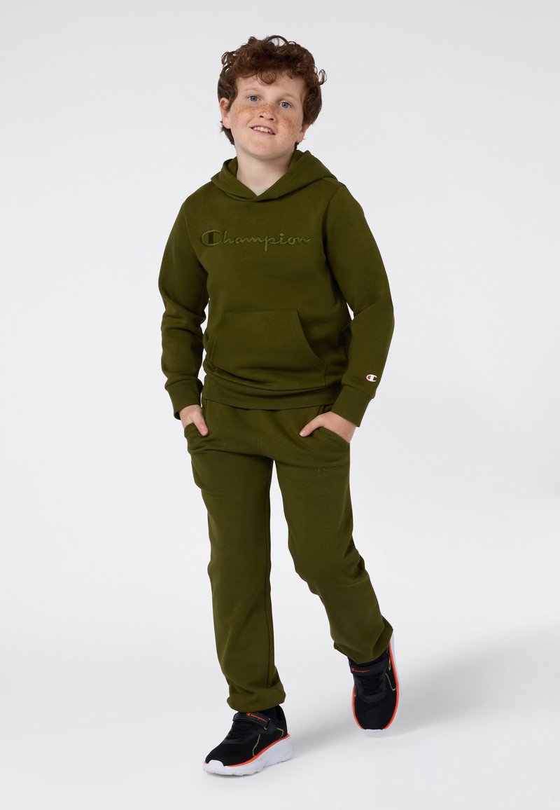 Felpa con cappuccio in cotone verde oliva con logo "Champion" sul petto, abbinata a pantaloni della tuta verdi oliva. Sneakers nere con accenti rossi e verdi.